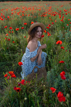 Young woman wearing straw hat smells red Papaver rhoeas flower in peaceful summer meadow landscape during sunset