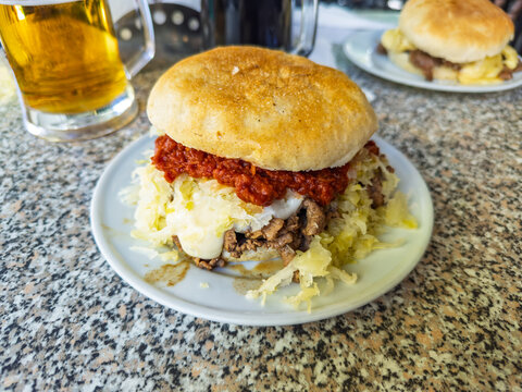 Chilean-style Churrasco with Italian toppings: A stacked sandwich on a rustic bread roll with beef, mayo, sauerkraut, and tomato-based sauce, served with a bee