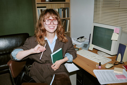 Portrait of smiling young adult Caucasian woman sitting at desk in retro office holding notebook and pencil. Image represents vintage corporate style, suitable for nostalgic business marketing