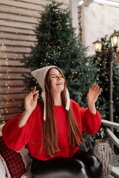 Laughing young woman wearing red sweater and white hat celebrates winter holidays outdoors near decorated fir trees