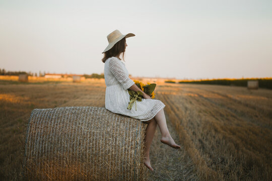 woman in a white dress holding a bouquet of wild flowers with sunflowers in a mowed field with straw bales