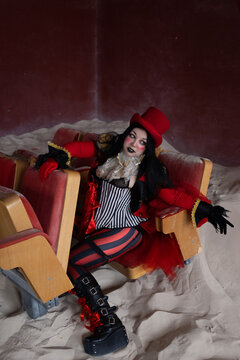 Reclining performer in red hat draped across rows of derelict theatre chairs in sand