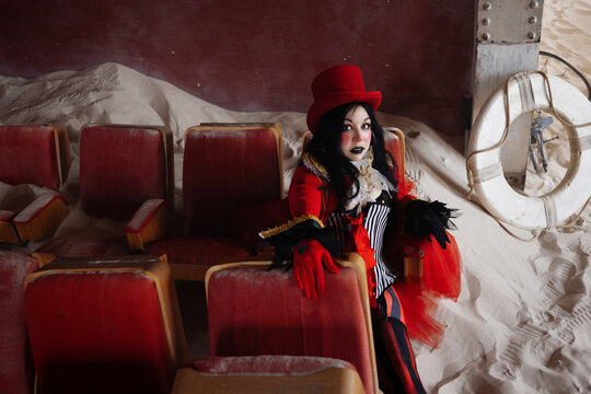 Worn red chairs, drifted sand and a rusted wheel leaning nearby frame the seated performer