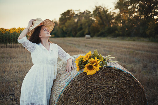 woman in a white dress holding a bouquet of wild flowers with sunflowers in a mowed field with straw bales