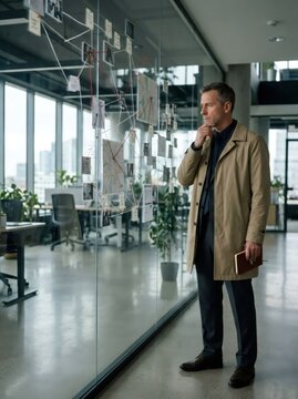 Thoughtful detective man in trench coat analyzing evidence board with photos, maps and notes connected by strings on glass wall in modern