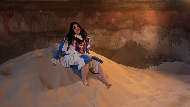 Barefoot woman in blue theatrical dress sitting on sand dune in dim abandoned hall