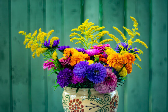 A bouquet of bright autumn flowers in a hand-painted ceramic vase against a green wooden wall.