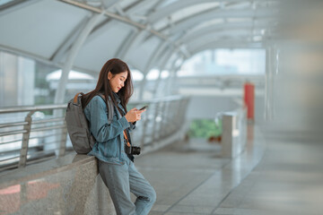 Young female traveler using smartphone with camera and backpack in modern public walkway,...