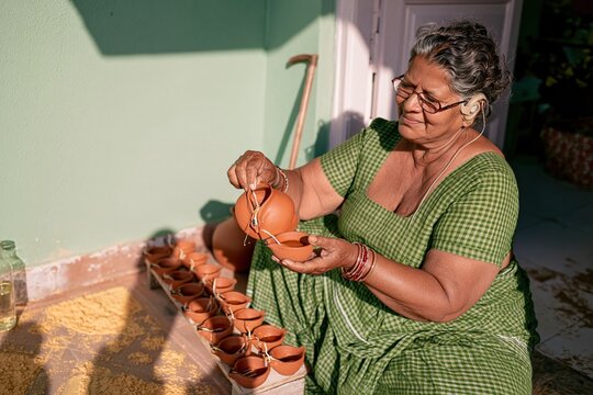 Senior Marwari woman filling terracotta oil lamps by a muted green wall, a natural scene of festival preparation for cultural and lifestyle content