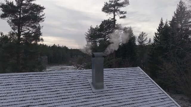 Smoke Billows From The Chimney Of A Cabin With A Snow-covered Roof In A Winter Landscape In Norway. Aerial Ascending Shot