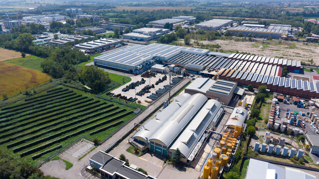 Aerial view of an industrial zone featuring chemical manufacturing and production facilities with solar panels on rooftops, surrounded by green fields in Cornaredo, Lombardy, Italy.