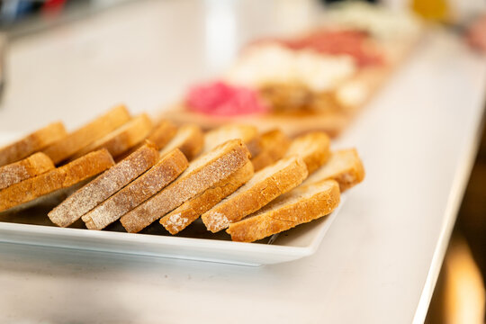 Toasted, grilled bread slices in rows on a white plate beside a charcuterie board at a bar, crisp golden crostini bases ready for cheese and cured meats.