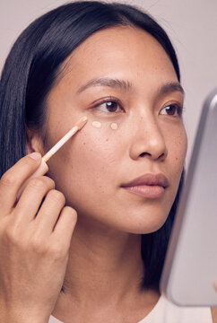 Closeup asian woman applying concealer under eyes with makeup wand while looking in mirror for beauty tutorial and cosmetic background