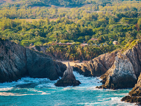 Aerial view of the Dedo de Dios rock formation rising from the turquoise sea, with a sandy beach, dense palm trees, and lush green hills in the background in Maruata, Michoacan, Mexico.