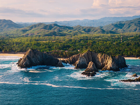 Aerial view of the Dedo de Dios rock formation surrounded by turquoise ocean waves and lush green coastal vegetation in Maruata, Michoacan, Mexico.
