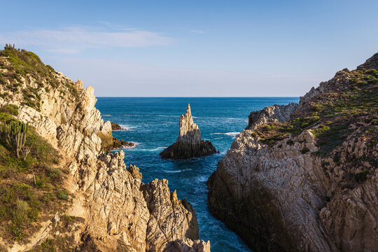 Aerial view of the Dedo de Dios rock formation rising from the blue ocean, surrounded by rugged coastal cliffs and lush vegetation in Maruata, Michoacan, Mexico.