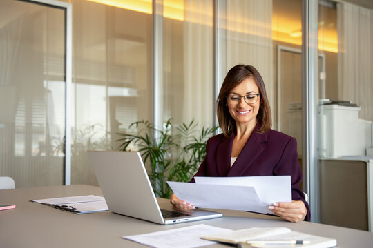 Mature businesswoman reading document, reviewing paperwork at office desk. Mid age business woman works as manager director executive, verifying client contract, processing vendor payment. Copy space.