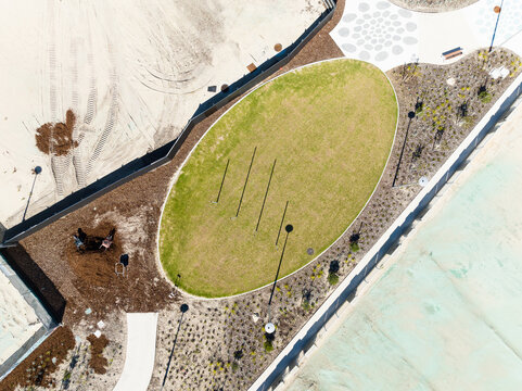 Aerial view of the landscaped oval lawn at Ocean Reef Boat Harbour, featuring modern paving, garden beds, and shadows cast by light poles in Ocean Reef, Western Australia, Australia.