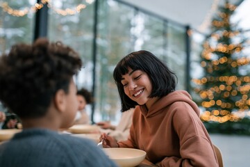 Fototapeta premium Young woman smiling across a table at a festive holiday meal with christmas tree and twinkling fairy lights for holiday greeting card and seasonal family card