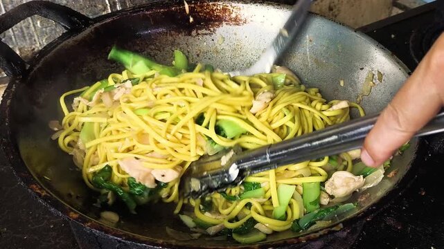 Close-up of chef hands using spatula to stir-fry yellow egg noodles with sliced chicken breast and fresh green bok choy in a traditional hot wok.  