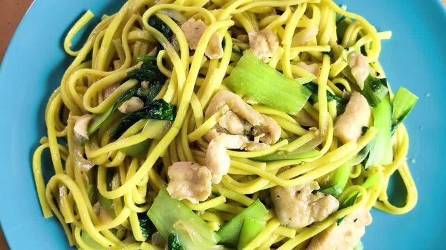 Top-down view of tasty stir-fried yellow egg noodles with tender chicken pieces and green bok choy rotating on a bright blue plate.