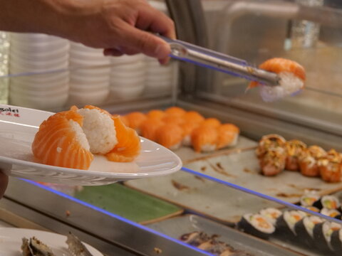 Customer taking salmon nigiri sushi with tongs from buffet display line in Japanese restaurant.