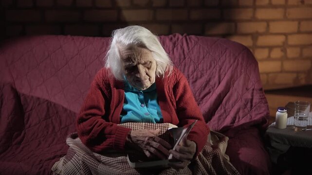 Old gray hair woman sits on a sofa in the room, holds a book and turns its pages