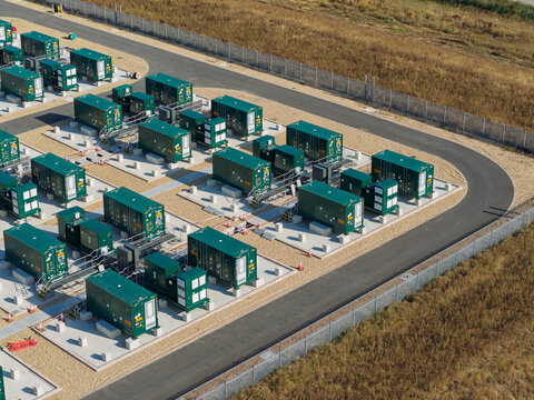 Aerial view of a power generation facility featuring rows of green containerized generators arranged on gravel pads with paved pathways and perimeter fencing in the United Kingdom.