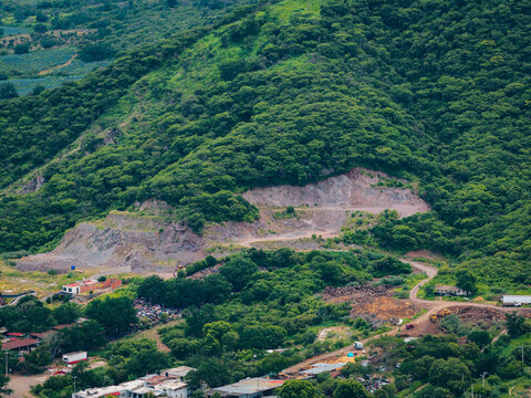 Aerial view of a quarry carved into a lush green hillside with exposed rock faces and dirt paths, overlooking a small town in Tuxpan, Jalisco, Mexico.