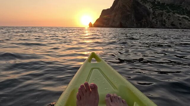 Sailing yellow kayak past rocky coastline during vibrant sunset. First-person POV wide shot.