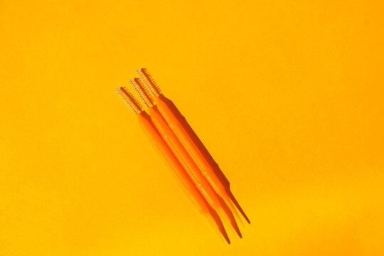 Three orange dental tools with pointed tips and bristles arranged on a bright yellow background for dental hygiene and oral care purposes