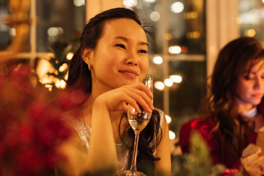 Asian woman holding a champagne glass smiles during a festive gathering with blurred lights and decorations in the background, another woman seated nearby