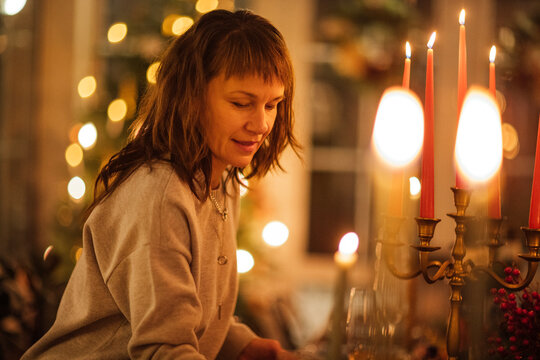 Woman with brown hair in gray sweater sets a festive table with candles and decorative elements, warm lighting creating a cozy atmosphere in a holiday setting