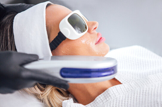 Female patient receiving facial treatment with laser device, wearing protective eyewear, in a modern spa setting, showcasing advanced skincare technology and relaxation