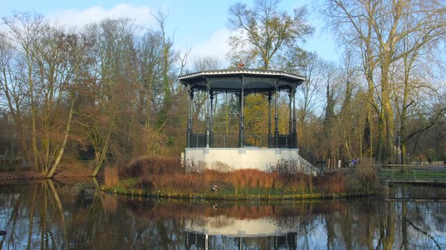 Scenic bandstand structure situated on small island within tranquil park pond during beautiful sunny autumn day with calm water reflections.