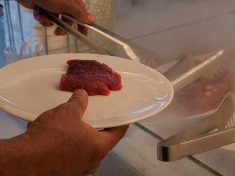 Customer holding white plate while picking raw red meat with metal tongs from a mist-cooled fresh food buffet station.