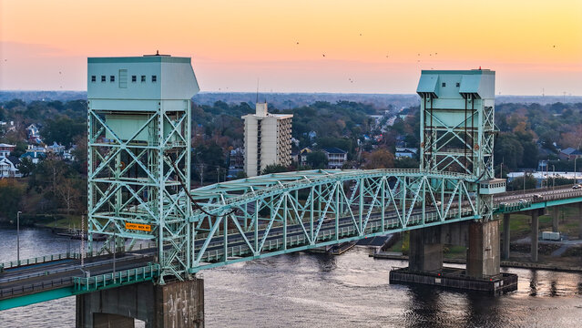 Aerial view of the Cape Fear Memorial Bridge spanning the Cape Fear River at sunset, with the city skyline and surrounding greenery in Wilmington, North Carolina, United States.