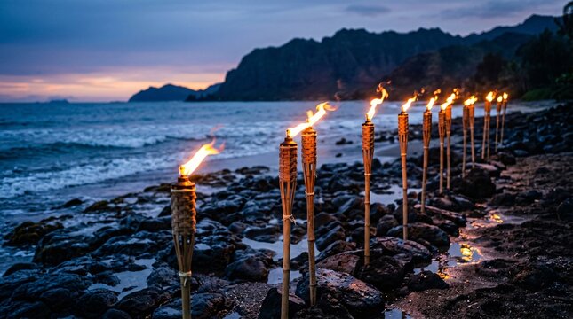 Burning bamboo tiki torches lined on dark rocky beach, vibrant orange flames against cool blue twilight ocean and distant mountains.