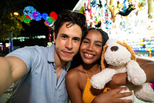 Diverse couple smiling, holding a stuffed animal, and taking a selfie at a night fair