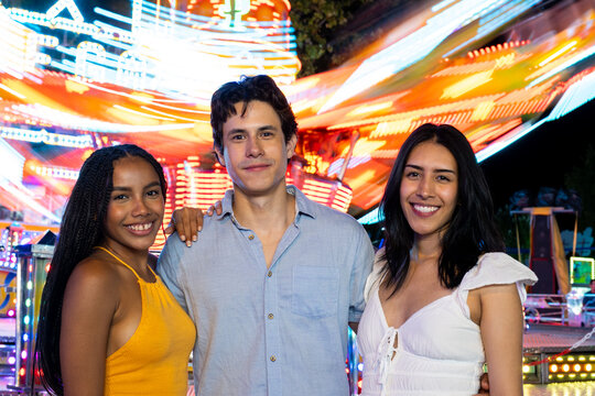 Three friends smiling and enjoying time together at a vibrant fairground at night