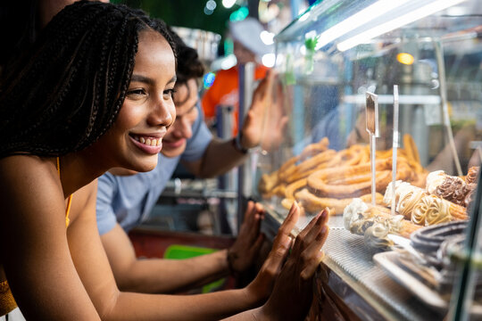 Young woman smiling while looking at churros in a food stall with a man behind her