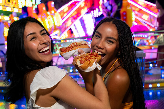Two cheerful friends eating hot dogs with neon lights blurring in the background