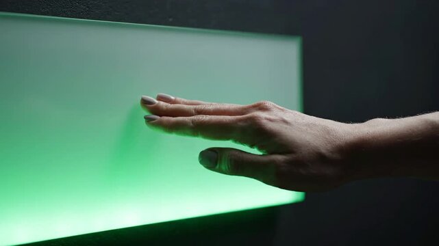 A hand interacts with a glowing white light panel. Upon contact, a bright spark occurs, and the surface transitions to a vivid green glow before the hand eventually moves away.