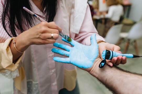 A professional artist uses a brush to paint and color the palm of a man's hand with blue watercolor paints to create an imprint.