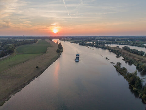 Aerial view of a cargo barge navigating a calm river during a golden sunset, surrounded by green fields and trees in The Netherlands.
