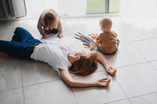 Tired mother lying on floor while her children drawing nearby.