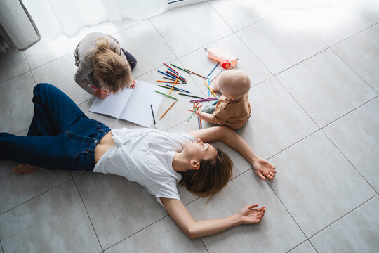Tired mother lying on floor while her children drawing nearby.