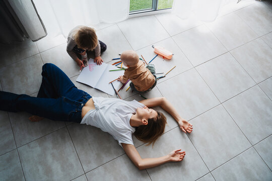 Tired mother lying on floor while her children drawing nearby.