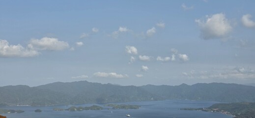 Naklejka premium Panoramic aerial view of calm blue sea, small green islands, and hazy mountains under a cloudy sky.