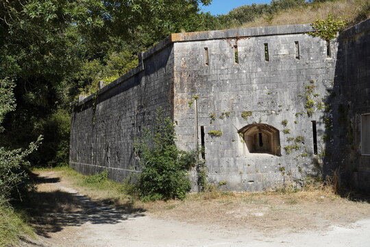 A massive stone wall of Fort Li&eacute;dot featuring narrow embrasures and embrasure slits stands surrounded by green foliage along a dirt path under direct sunlight.  